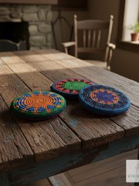 Three colorful, round crocheted coin purses sit on a rustic wooden table in a cozy room with a stone fireplace, wooden chair, and sunny window. The crocheted coin purses have vibrant patterns and the room has a warm, inviting atmosphere.