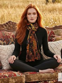 A woman with long red hair sits cross-legged on a vintage patterned sofa outdoors, wearing a black outfit and a multicolored scarf. The setting is a field with tall grass, creating a bohemian, autumn atmosphere.