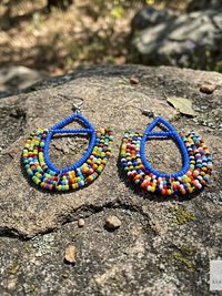 A pair of colorful beaded earrings with blue outlines and multicolored beads, displayed on a large rock outdoors with a blurred natural background.