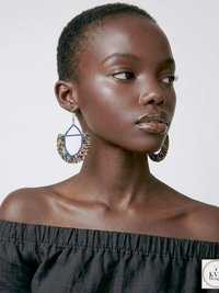 A woman with short hair wears large, colorful beaded hoop earrings and an off-the-shoulder black top, posing against a plain light background.