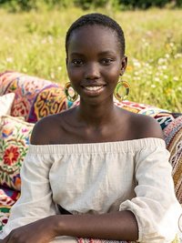 A woman with short hair and large hoop beaded maasai earrings sits on a colorful, patterned couch in a sunny, grassy field, smiling at the camera. She wears an off-the-shoulder, light-colored dress.