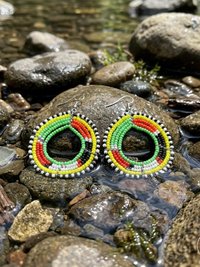 A pair of colorful beaded hoop earrings with red, green, yellow, black, and white beads displayed on wet rocks in a shallow stream.