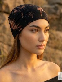 A woman with long hair wearing a black and brown tie-dye headscarf gazes into the distance outdoors, with sunlight illuminating her face. Rocks and warm natural light create a serene, earthy atmosphere.