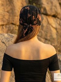 A person with long hair wears an off-shoulder black top and a black and brown headscarf, standing outdoors with rocky terrain in the background.