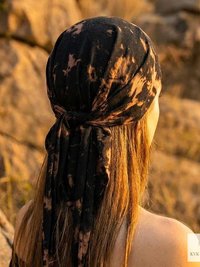 A person with long hair, seen from behind, wears a black and tan tie-dye headscarf outdoors near rocks. Warm sunlight highlights their hair.