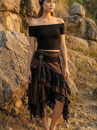 A woman in a black off-shoulder top, layered brown skirt, and headscarf stands barefoot on rocky terrain, surrounded by large rocks and dry grass, with the sun casting a warm light.