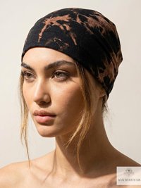 Woman with light makeup wears a black and brown tie-dye headwrap. She faces slightly left with a neutral expression. A few hair strands frame her face. The background is plain, and a KVK BOHOFAIR logo appears in the bottom right corner.