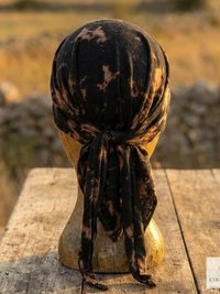 A black and brown tie-dye headscarf is tied on a mannequin head, placed on a rustic wooden table outdoors, with a blurred field and stone wall in the background.