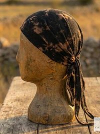 A wooden mannequin head displays a black and tan tie-dye headscarf, tied at the back, set outdoors on a wooden table with a blurred field and stone wall in the background.
