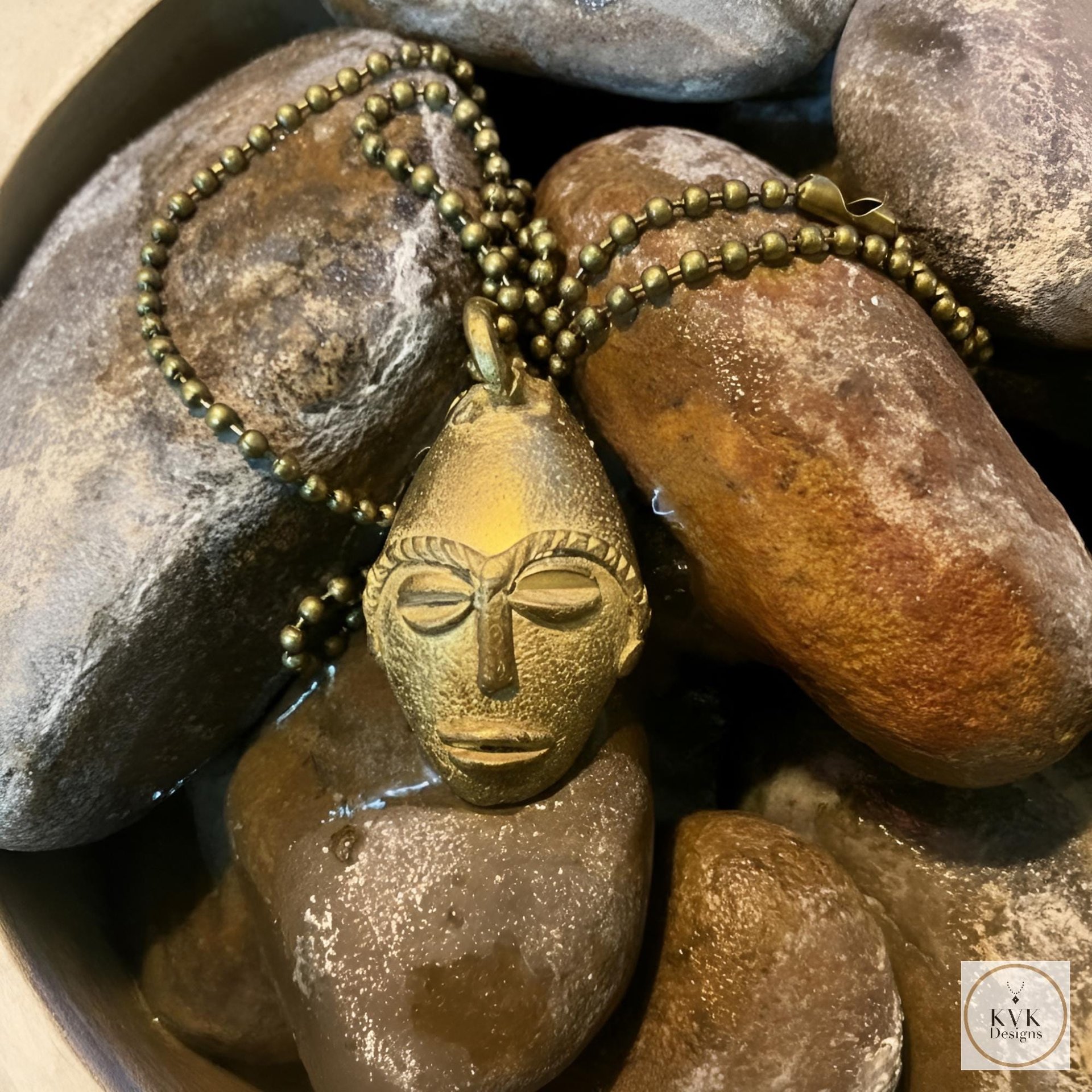 A brass pendant necklace with a stylized face design rests on smooth, wet stones. The pendant is attached to a beaded chain