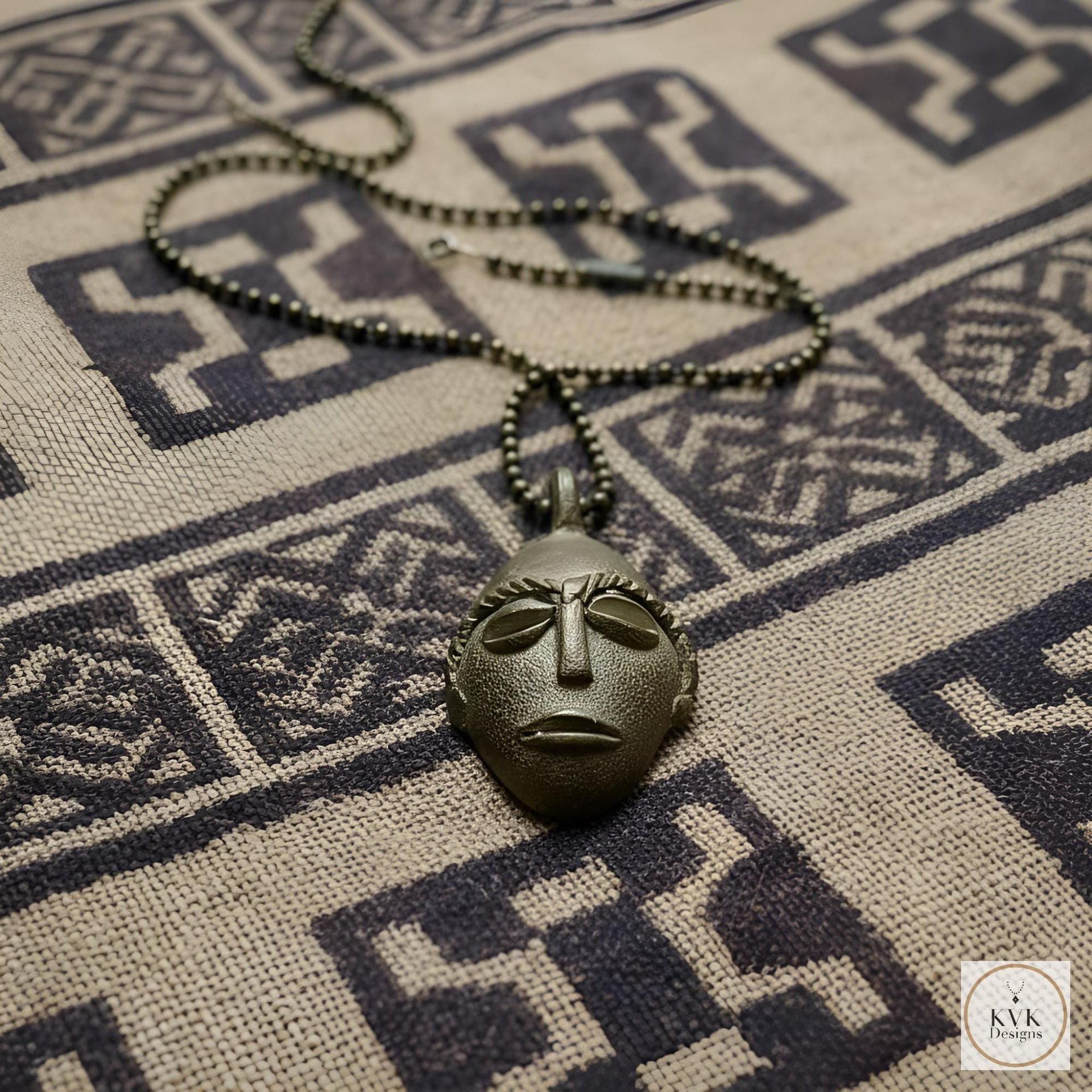 A metallic pendant shaped like an abstract face rests on a patterned fabric with geometric designs. The pendant hangs from a beaded chain.
