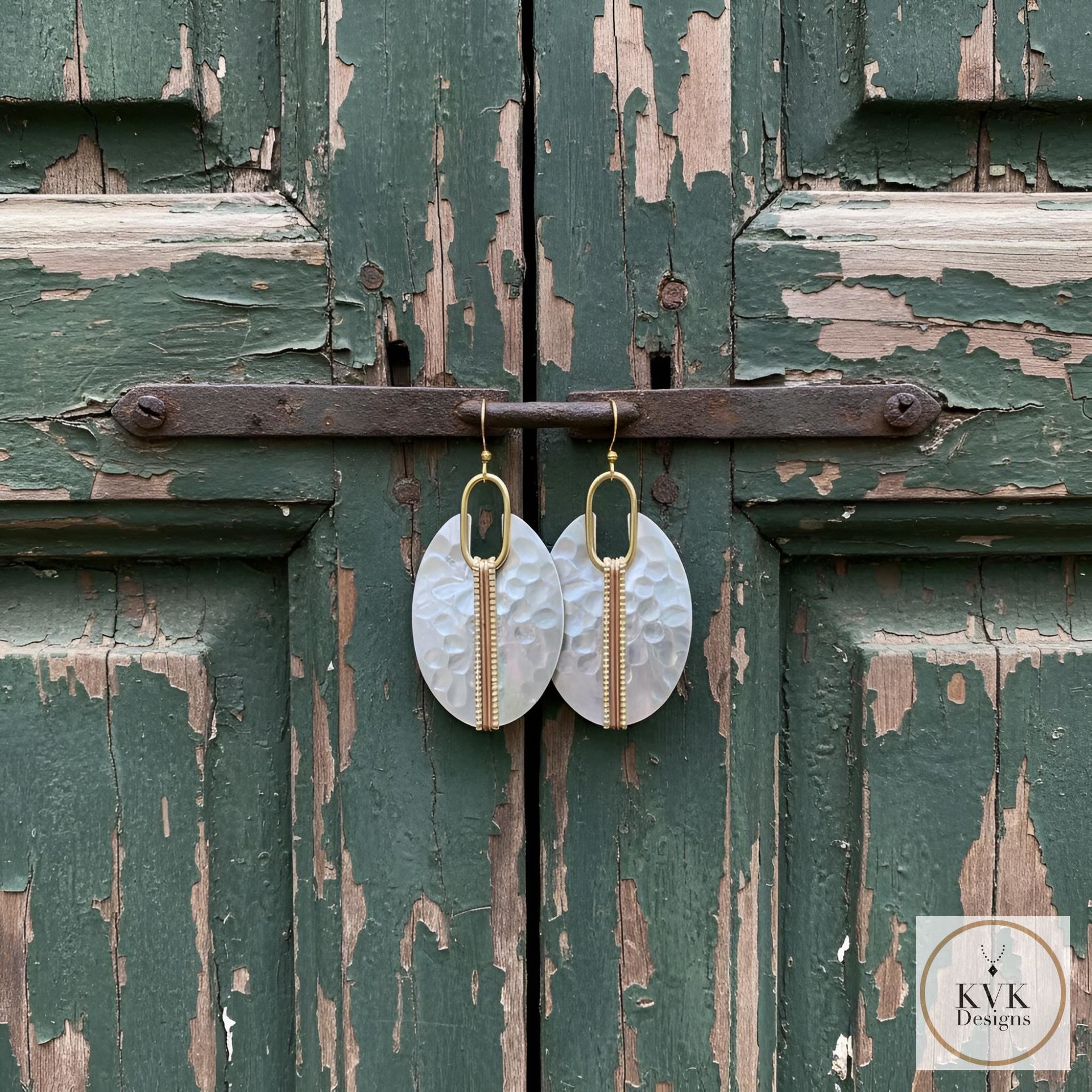 Oval-shaped earrings with a hammered texture and a gold-colored vertical accent. The earrings are suspended from gold-colored hooks. The background is a weathered green wooden door.