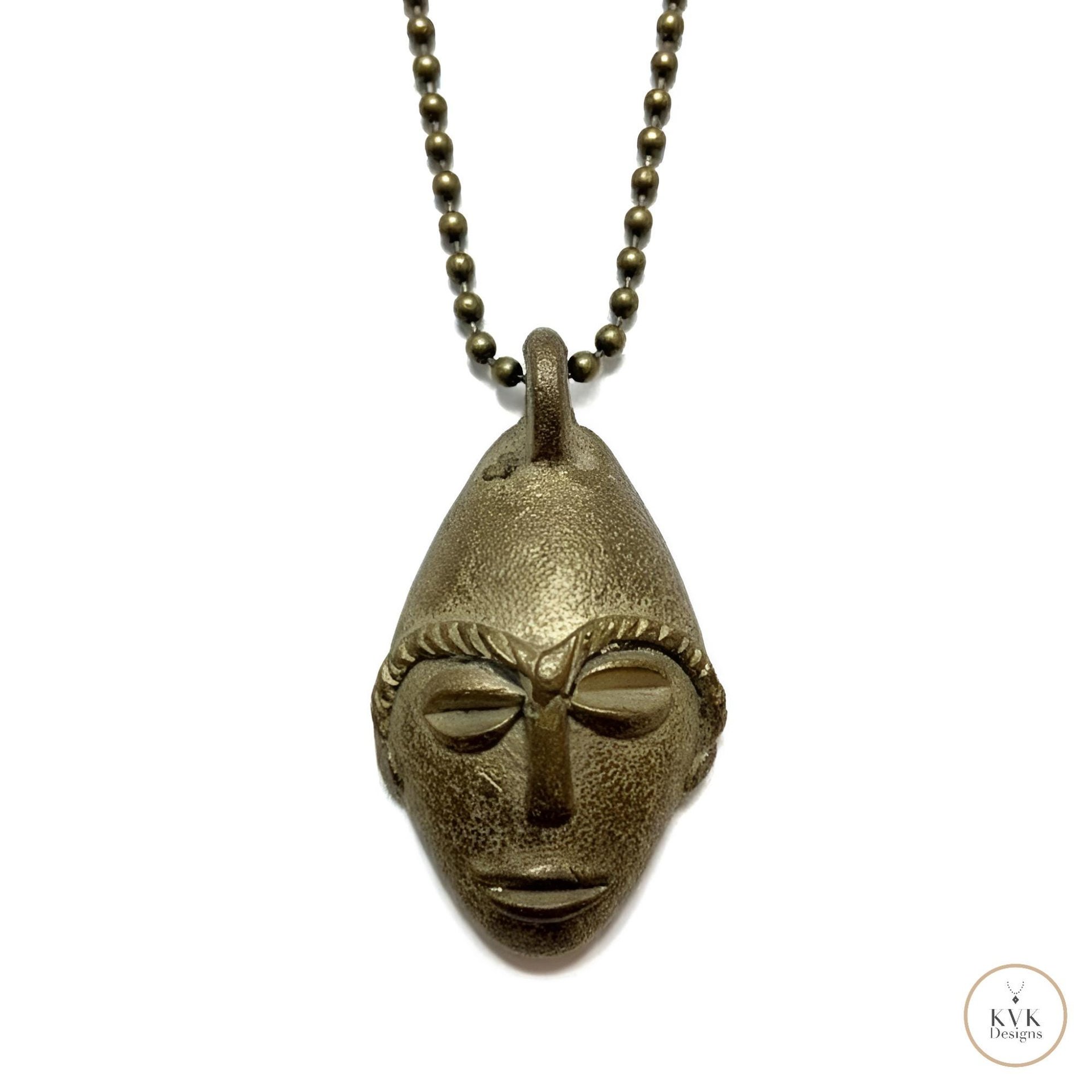 A brass pendant in the shape of a stylized human face with prominent brows, nose, and lips, hanging from a ball chain necklace on a white background.