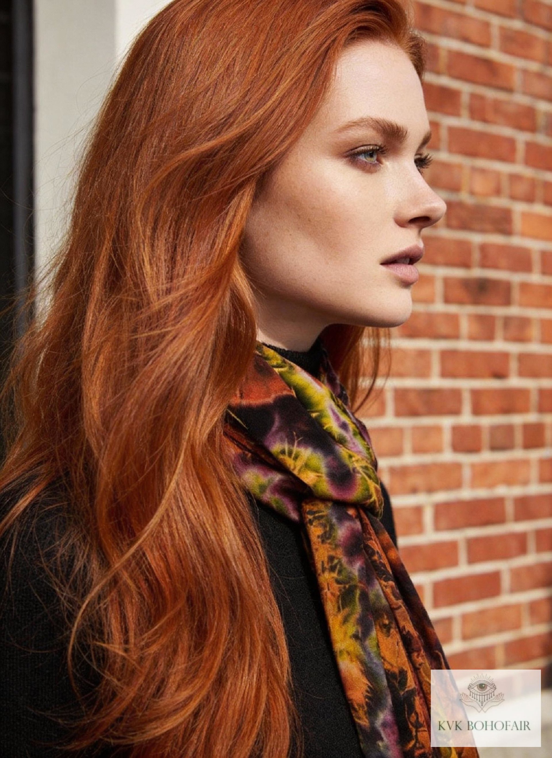 A woman with long red hair, wearing a colorful tie dye patterned scarf and a black top, stands in profile against a red brick wall.