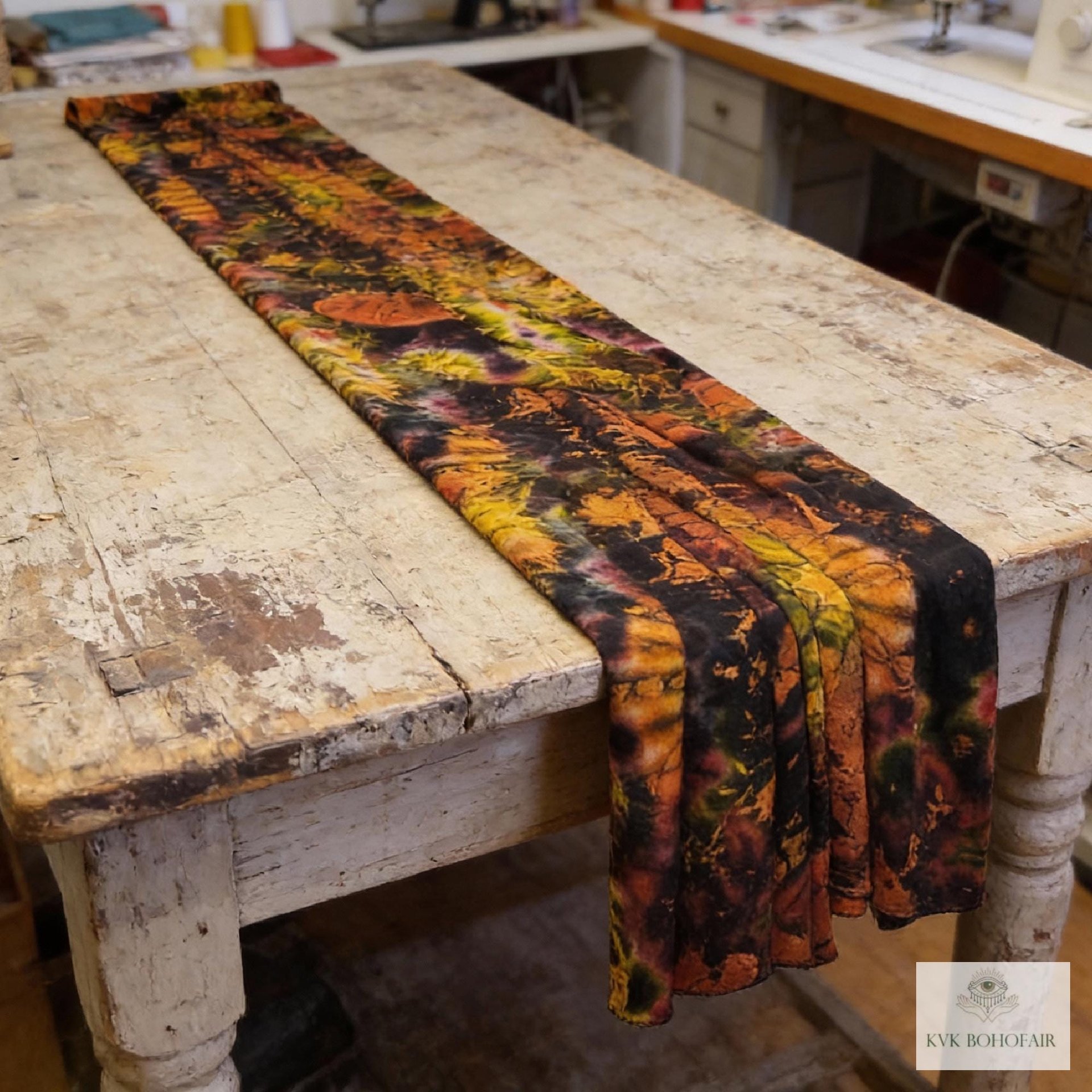 A long, rectangular scarf with a tie-dye pattern in shades of orange, yellow, and black. The scarf is draped across a distressed wooden table, showcasing the vibrant colors and unique design. The scarfs pattern creates a striking visual contrast.