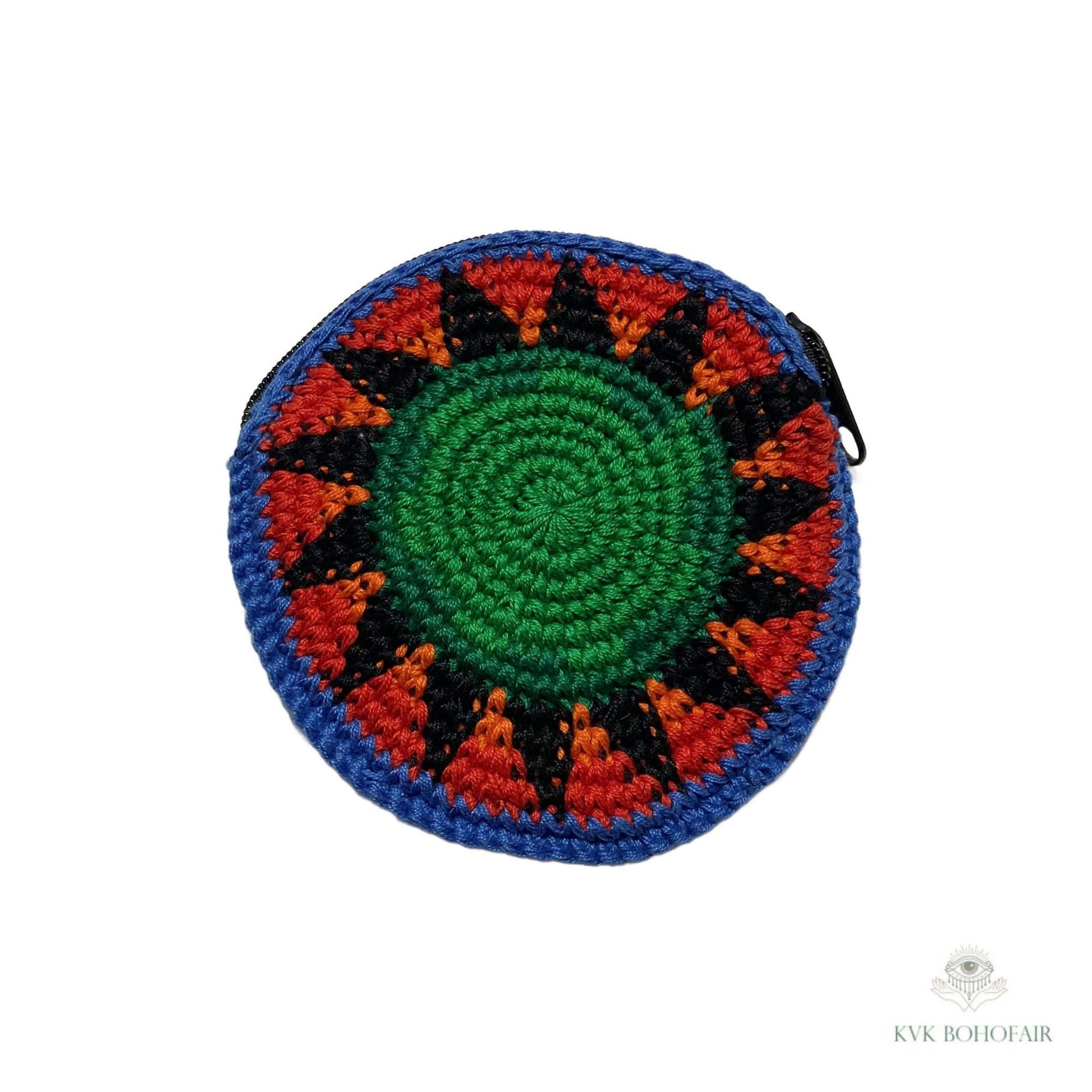 A round, crocheted coin pouch with a green center, surrounded by orange, black, and blue geometric patterns, and a zipper on one side. The background is white with a small watermark in the bottom right corner.