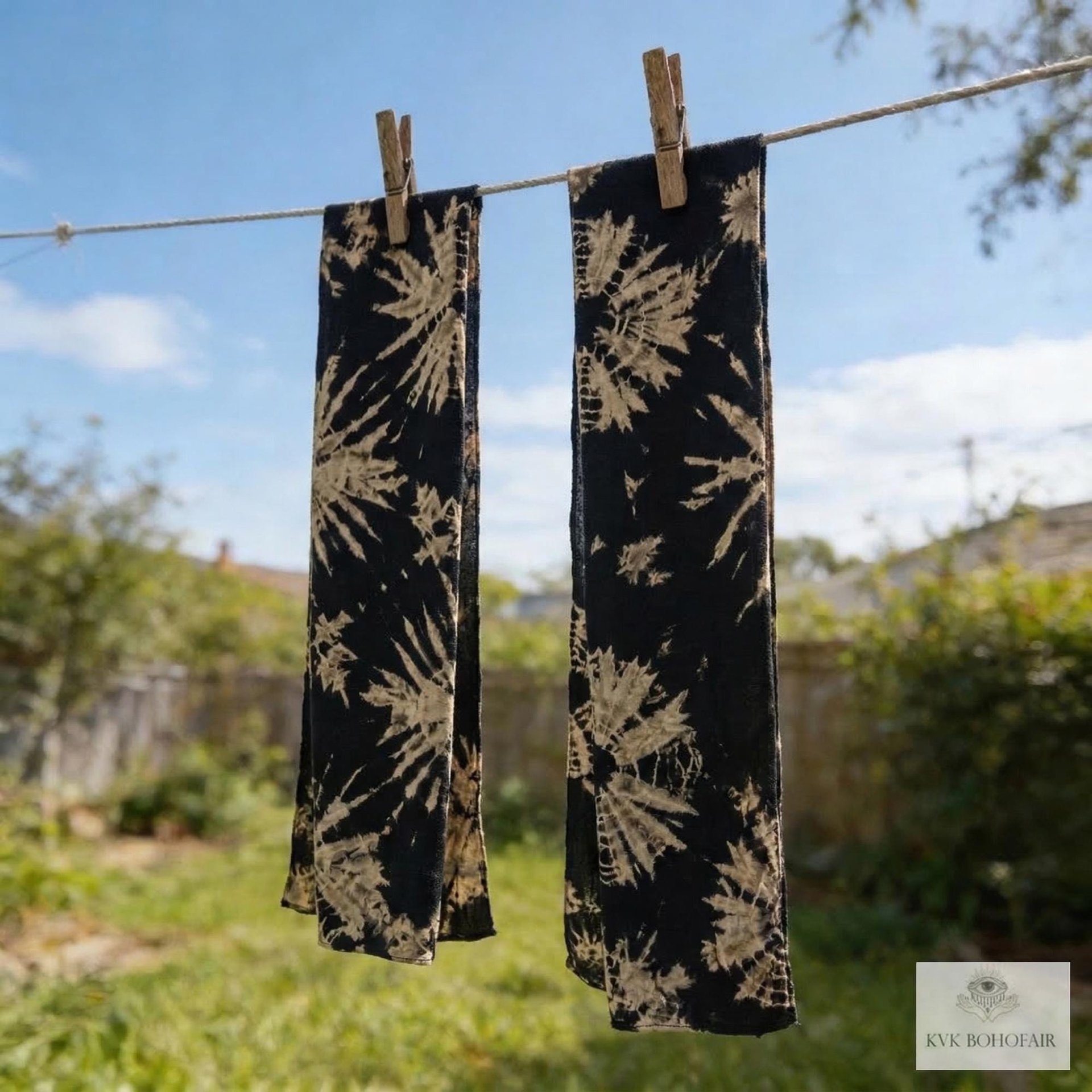 Two black and beige tie-dye scarves hang from a clothesline with wooden clothespins, drying outdoors in a sunny backyard with green grass, a fence, and trees in the background.