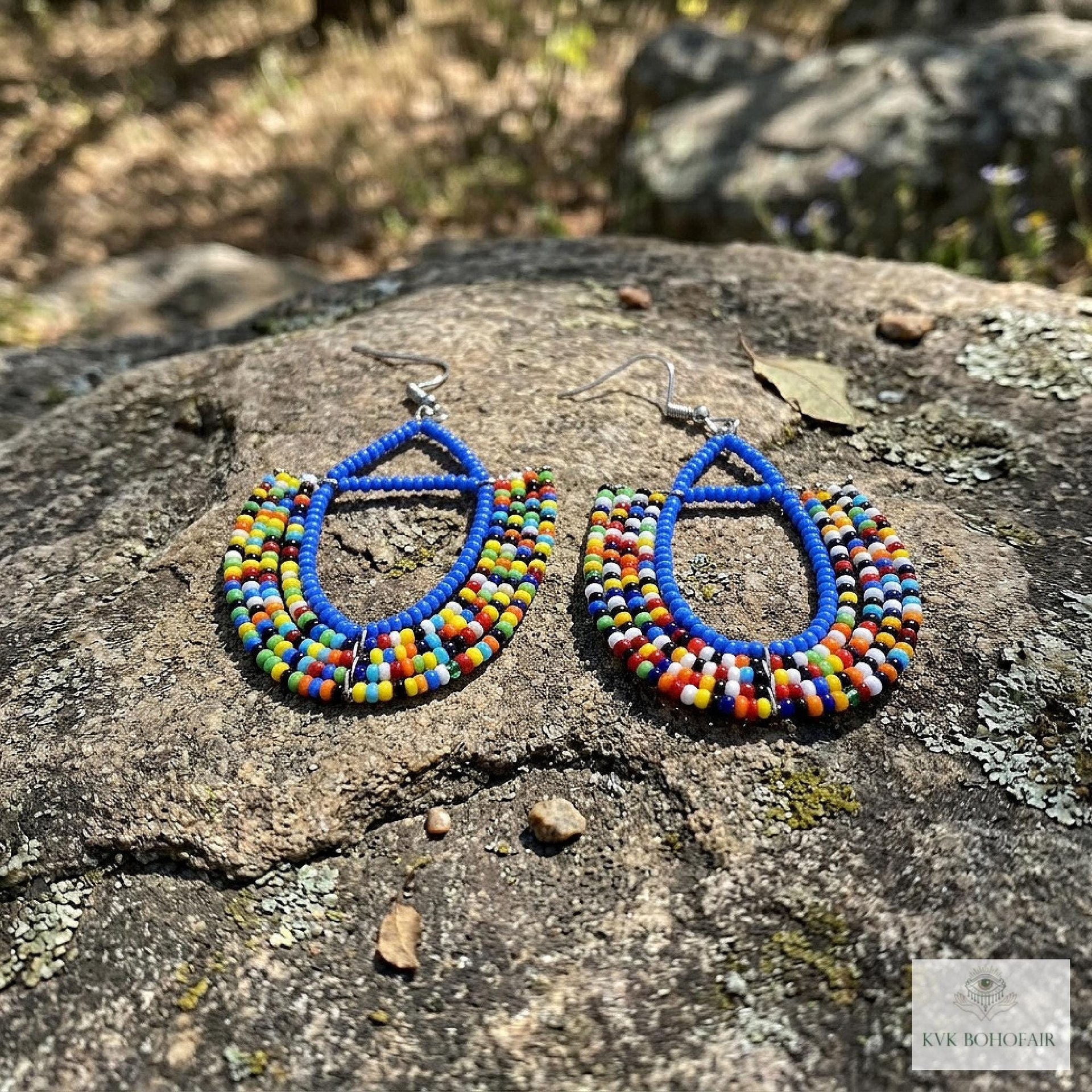 A pair of colorful beaded earrings with blue outlines and multicolored beads, displayed on a large rock outdoors with a blurred natural background.