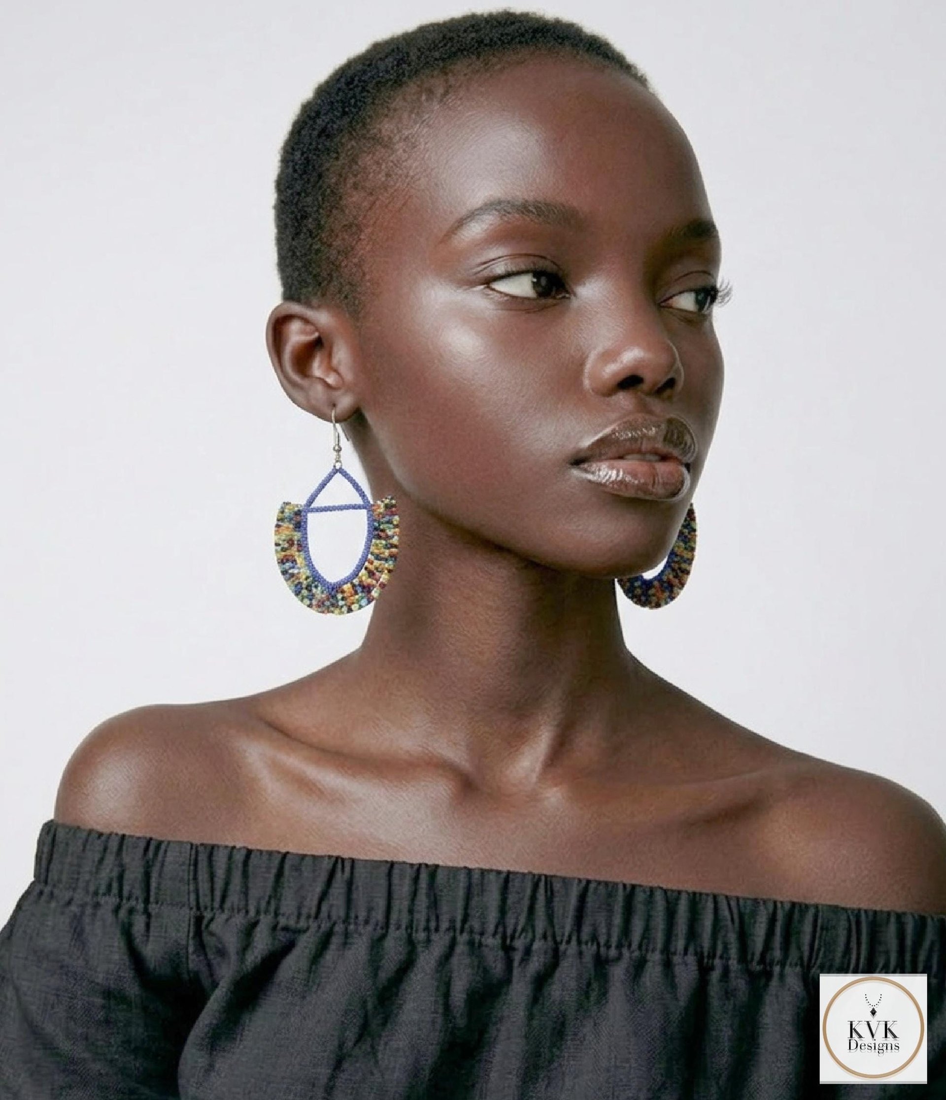 A woman with short hair wears large, colorful beaded hoop earrings and an off-the-shoulder black top, posing against a plain light background.