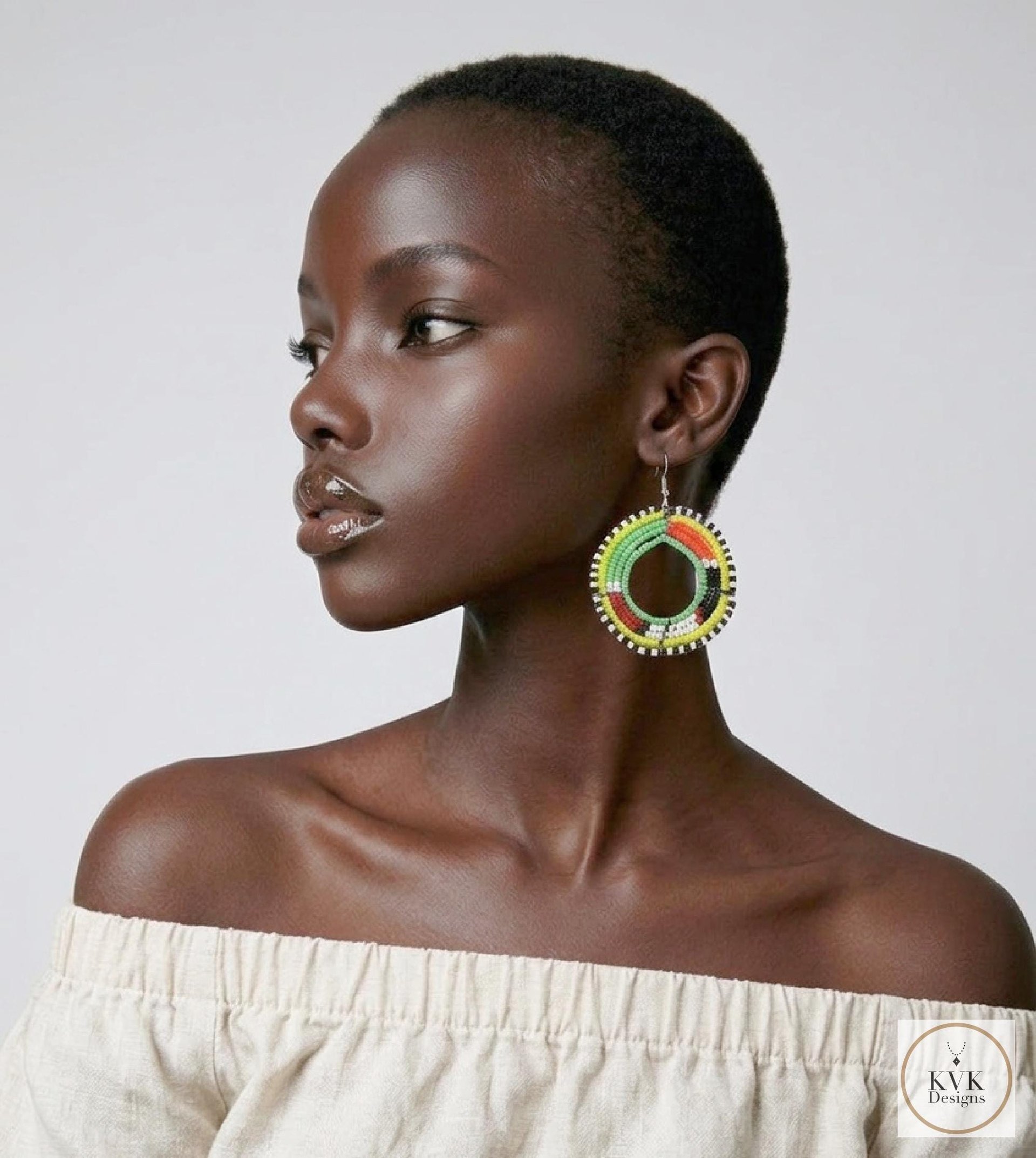 A woman with dark skin and short hair wears a cream off-shoulder top and colorful beaded hoop earrings. She looks to the side against a plain background.