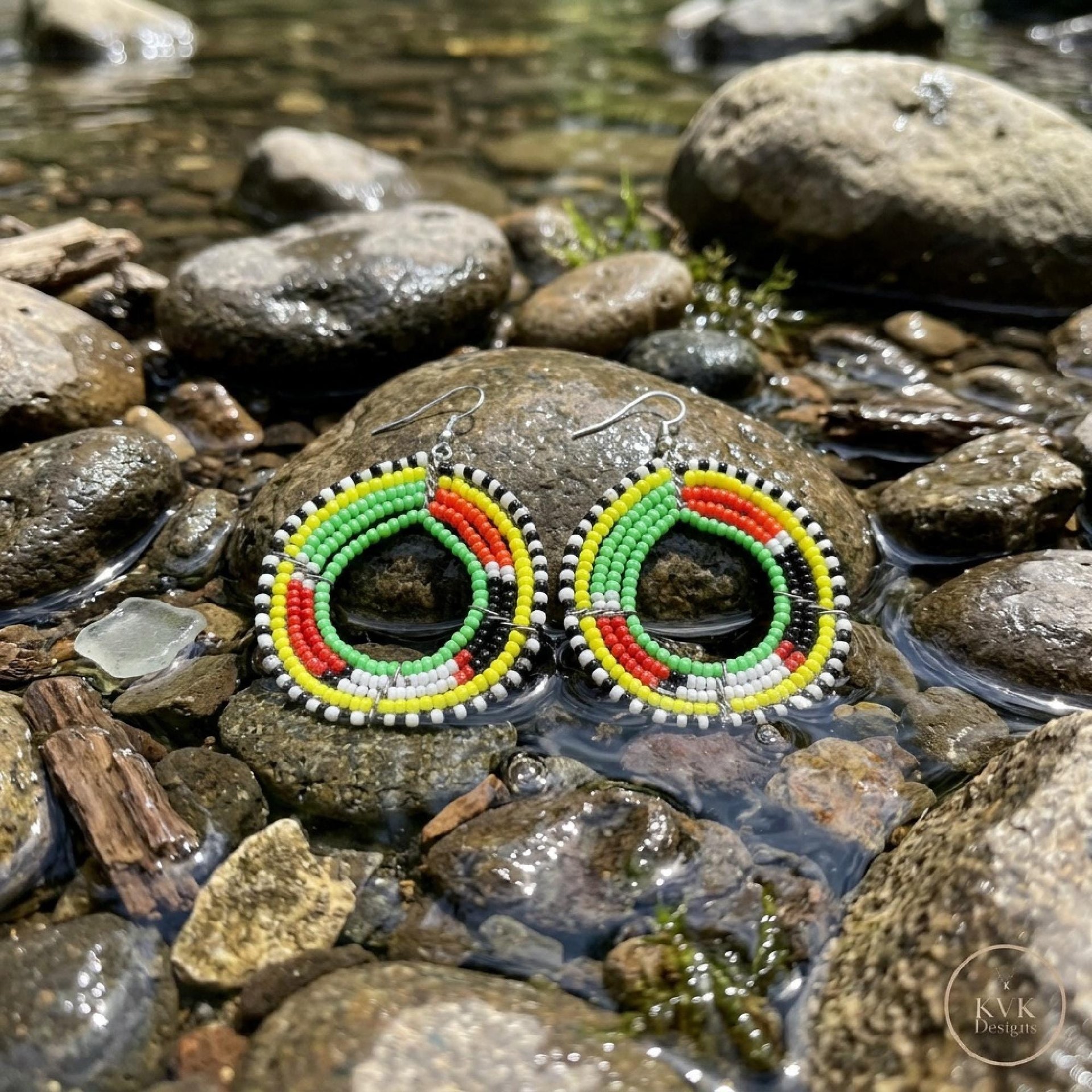 A pair of colorful beaded hoop earrings with red, green, yellow, black, and white beads displayed on wet rocks in a shallow stream.