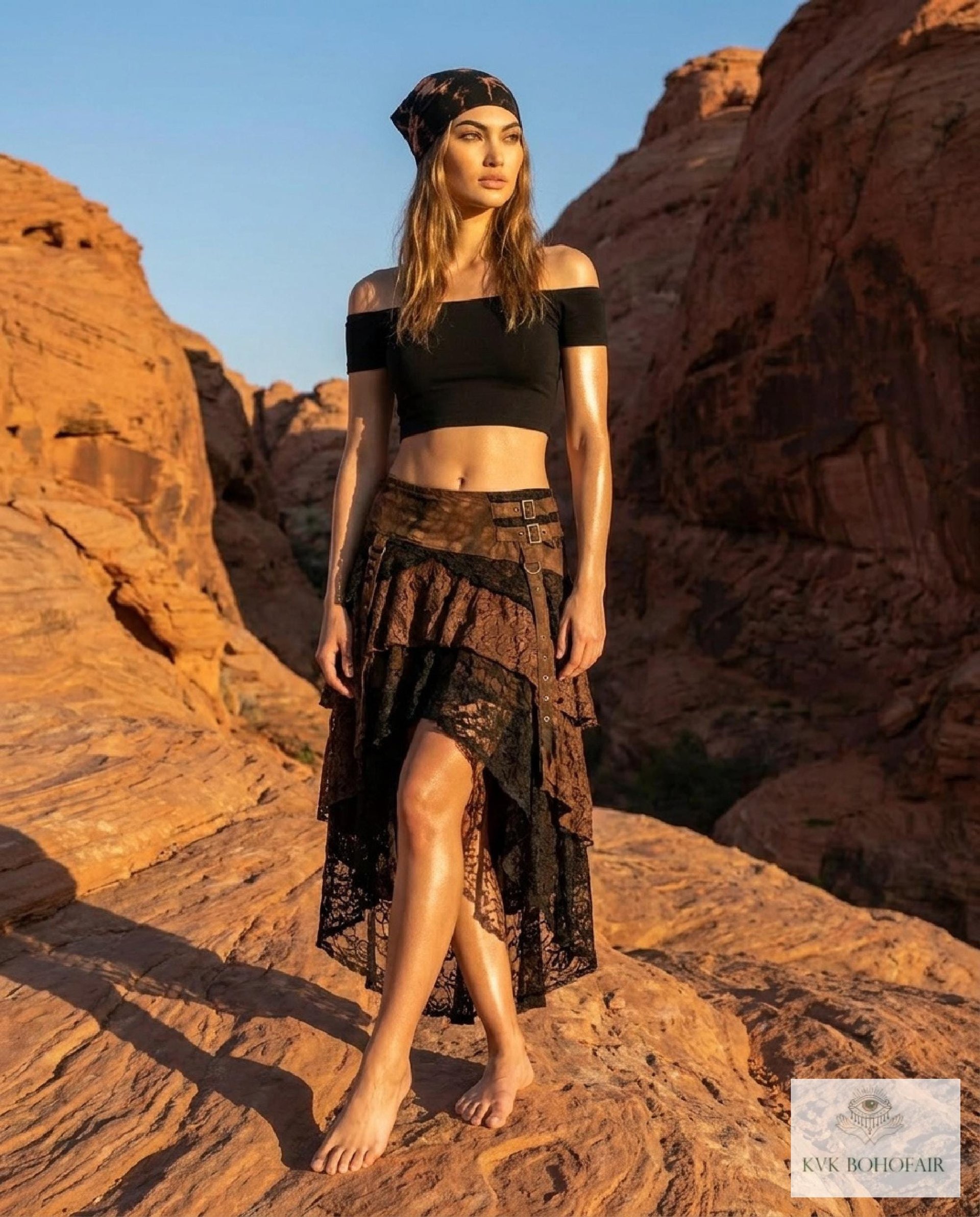 A woman in a black crop top, lace layered skirt, and headscarf stands barefoot on red rocks in a desert landscape, with sunlight highlighting her and rocky cliffs behind her.