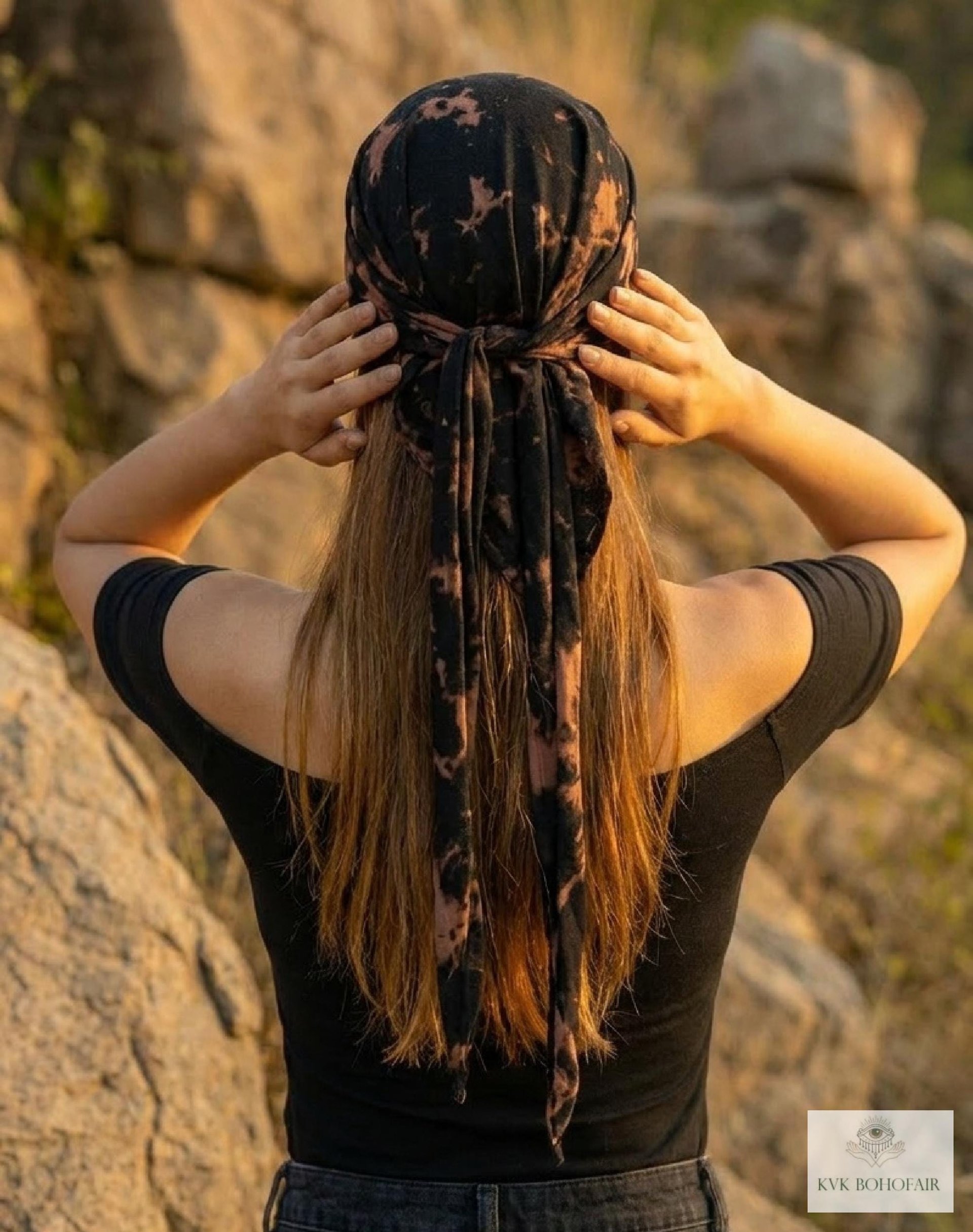 A woman with long hair, seen from behind, stands outdoors touching a black and brown tie-dye headscarf. She wears a black off-shoulder top and is surrounded by large rocks.