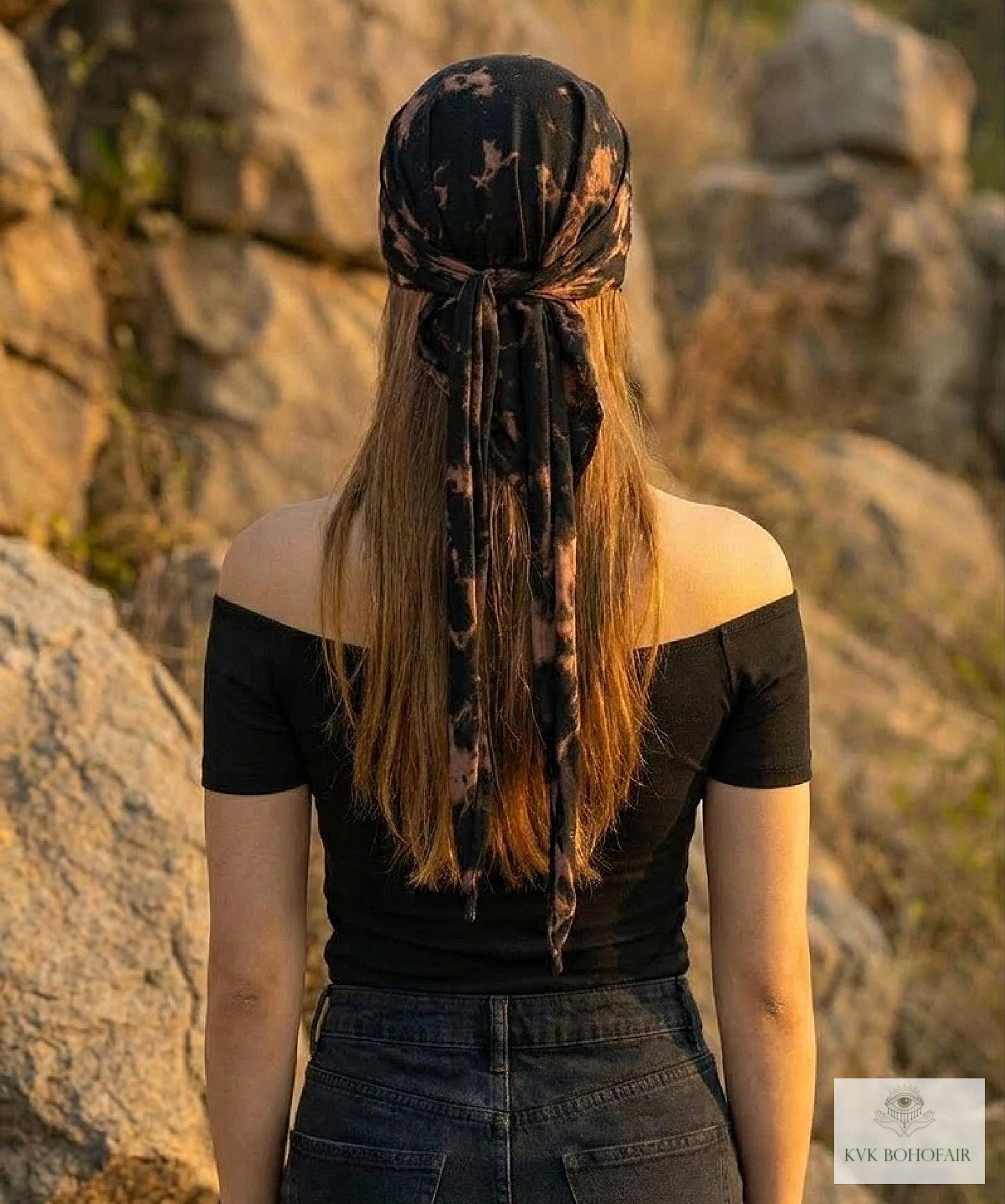 A person with long hair, wearing a black off-shoulder top and dark jeans, stands outdoors facing away. They have a black patterned scarf tied around their head. Rocks and greenery are in the background.