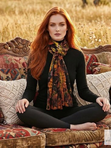 A woman with long red hair sits cross-legged on a vintage patterned sofa outdoors, wearing a black outfit and a multicolored scarf. The setting is a field with tall grass, creating a bohemian, autumn atmosphere.
